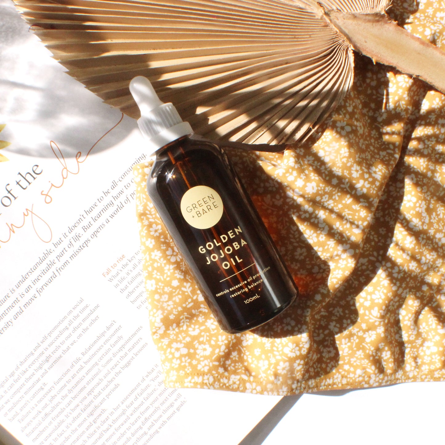 Flatlay image with palm leaf fan, golden jojoba brown glass bottle and floral dress